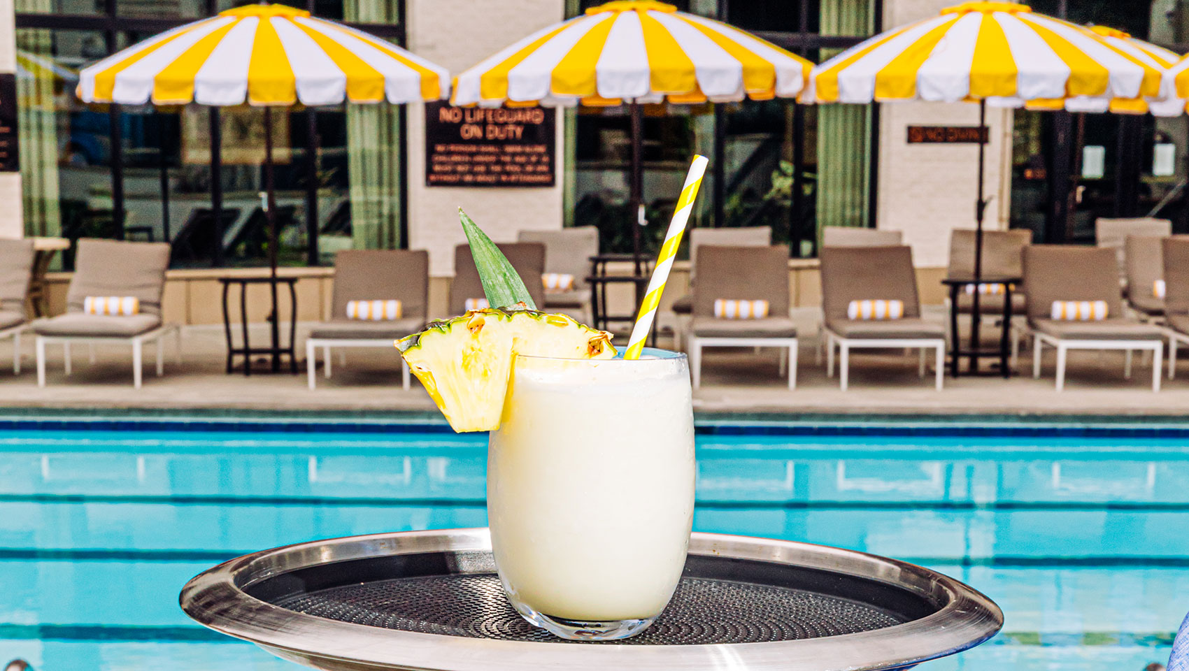 Hand holding a tray with a cocktail on it in front of the Kimpton Cottonwood Hotel Pool